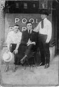 This is one of the earliest photographs of Al Capone. It was taken the day he graduated high school and before he had the scars on his face. He is sitting in front of the pool hall where his father, sitting next to Al, died of a massive heart attack in 1920. Look at the reflection in the window and you will see the apartment building across the street where they lived. The woman standing on the balcony is Al’s mother Theresa holding my father who was just a month old. The man standing is Vincenzo Raiola. 1917.