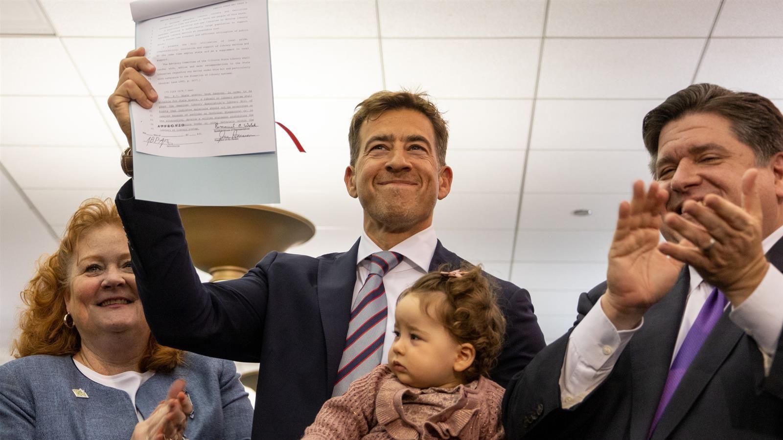 Illinois Secretary of State Alexi Giannoulias holds his daughter while brandishing a bill signed by Gov. J.B. Pritzker on June 12, 2023, that aims to discourage book bans at Illinois libraries. Bill sponsor Sen. Laura Murphy, D-Des Plaines, is pictured at left and Pritzker is pictured at right. (Capitol News Illinois photo by Andrew Adams)
